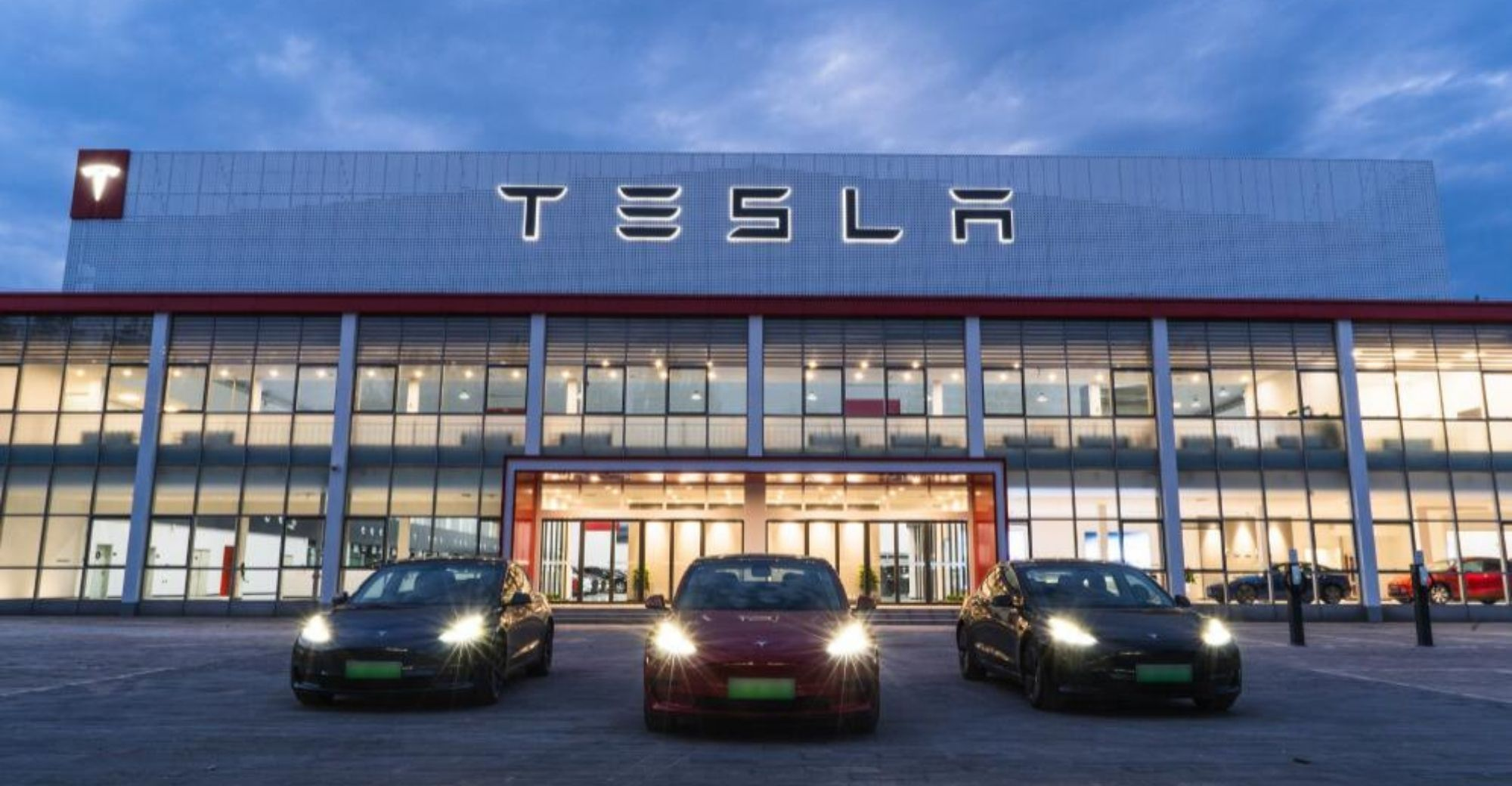 Tesla dealership showroom at dusk with three Tesla vehicles out front under an illuminated Tesla sign